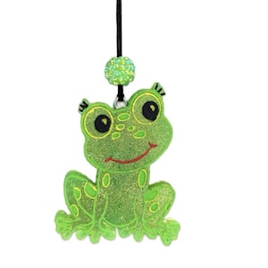May include: A green glittery frog-shaped pendant with a smiling face, large black eyes, and yellow accents. The frog is suspended from a black cord with a matching green glitter bead. The pendant is a whimsical accessory.