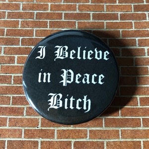 May include: A black button with white text that reads "I Believe in Peace Bitch" in a gothic font.