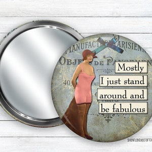 May include: A round compact mirror with a silver frame. The mirror's design features a vintage illustration of a woman in a pink dress with the text "Mostly I just stand around and be fabulous".