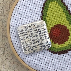 May include: A white fabric with a green and brown avocado design stitched into it. The fabric is stretched over a wooden embroidery hoop. A small white card with the text "DATE DUE" and a list of dates is attached to the fabric.