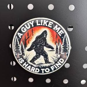 May include: Circular sticker with a Bigfoot illustration. The design features a black silhouette of Bigfoot walking in front of a forest scene with the text "A Guy Like Me Is Hard To Find" in a circular pattern.