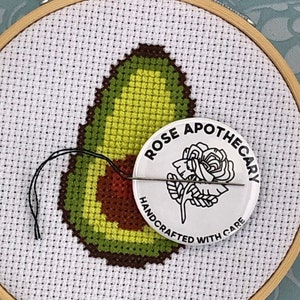 May include: A white embroidery hoop with a green and brown avocado cross-stitch design. A white button with black text reads "ROSE APOTHECARY HANDCRAFTED WITH CARE" and features a black line drawing of a rose.
