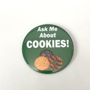 May include: A circular, green badge with the words "Ask Me About COOKIES!" in white. The badge shows illustrations of three cookies: a chocolate-covered biscuit, a shortbread biscuit, and a caramel coconut biscuit.