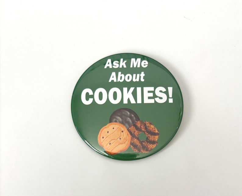 May include: A circular, green badge with the words "Ask Me About COOKIES!" in white. The badge shows illustrations of three cookies: a chocolate-covered biscuit, a shortbread biscuit, and a caramel coconut biscuit.