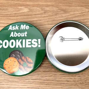 May include: Two round green buttons. One button has the text "Ask Me About COOKIES!" in white, with images of cookies. The other button is silver with a safety pin backing. The buttons are on a wooden surface.