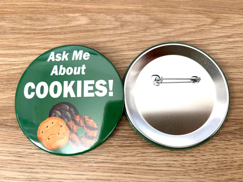 May include: Two round green badges. One badge has the text "Ask Me About COOKIES!" in white, with images of cookies. The other badge is silver with a safety pin backing. The badges are on a wooden surface.