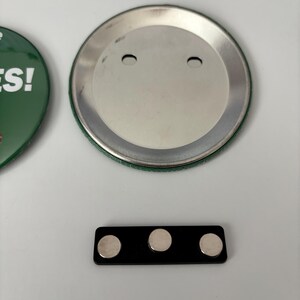 May include: A green button with the word "YES!" in white text, a silver button back, and a black magnetic bar with three silver magnets. The items are on a white surface.