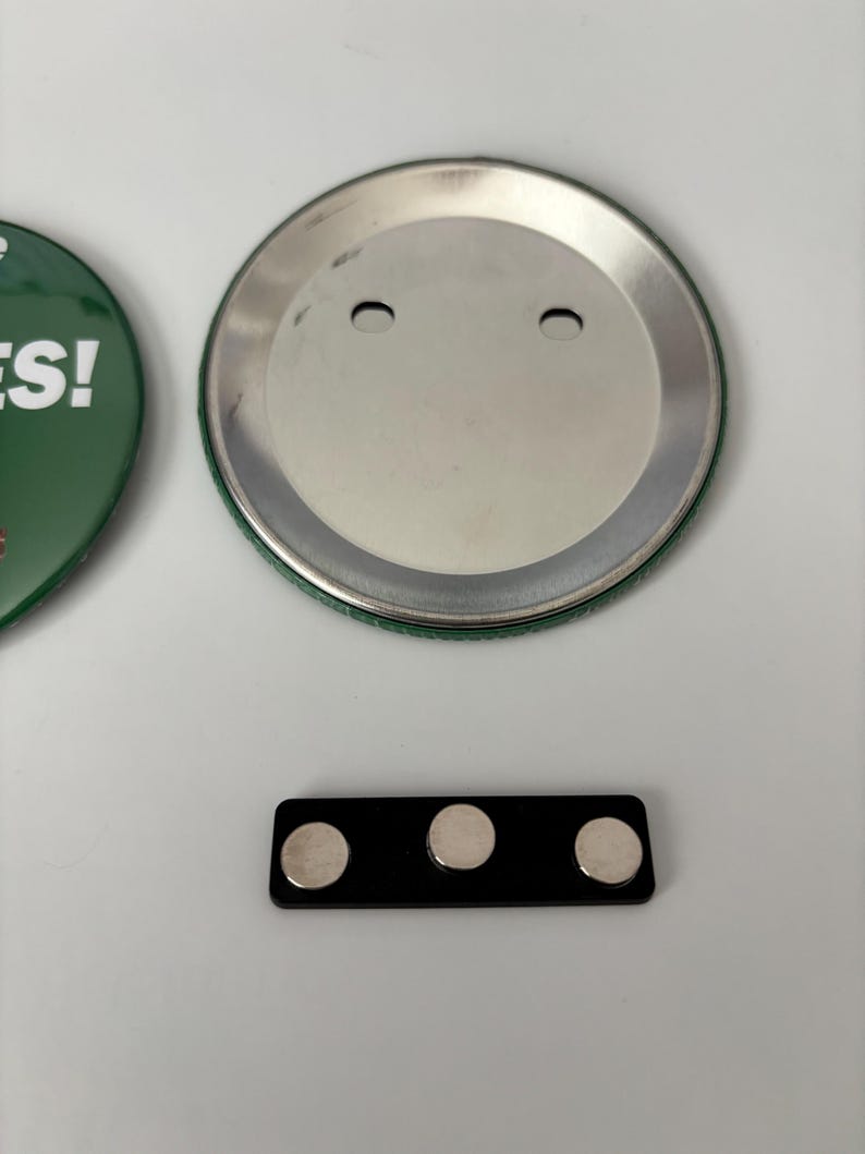 May include: A green button with the word "YES!" in white text, a silver button back, and a black magnetic bar with three silver magnets. The items are on a white surface.
