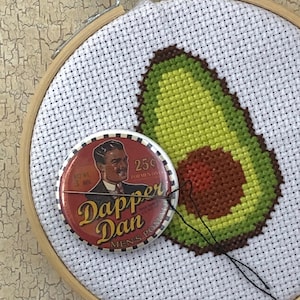 May include: A cross-stitch embroidery hoop with a green avocado design. A vintage Dapper Dan men's pomade tin is attached to the hoop.