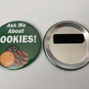 May include: A circular green magnet with the text "Ask Me About COOKIES!" in white. The magnet shows illustrations of three biscuits. The reverse side is silver with a black magnetic strip.