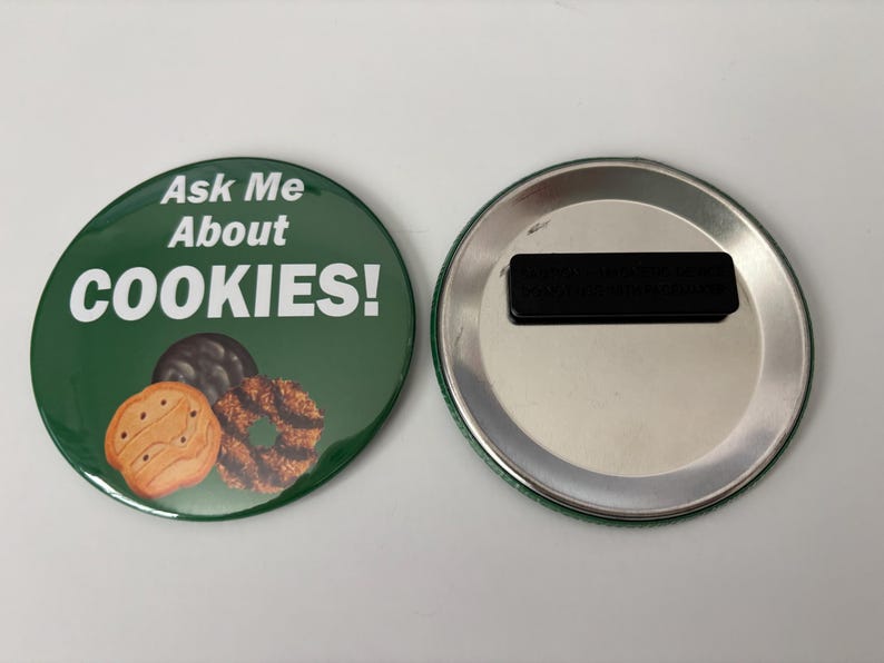 May include: A circular green magnet with the text "Ask Me About COOKIES!" in white. The magnet shows illustrations of three biscuits. The reverse side is silver with a black magnetic strip.