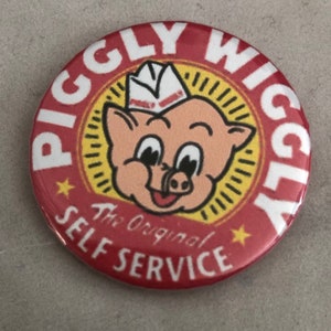 May include: Red and yellow button with a cartoon pig wearing a white paper hat. The text on the button reads "Piggly Wiggly The Original Self Service".