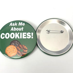 May include: A green badge with the words "Ask Me About COOKIES!" in white. The badge shows images of three biscuits. The reverse side of the badge is silver with a pin.