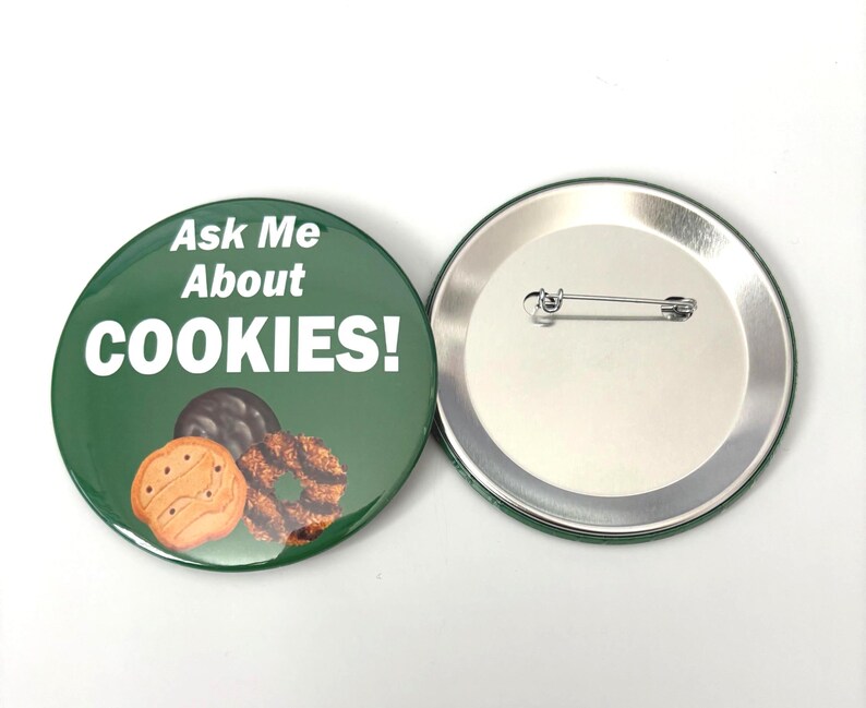 May include: A green badge with the words "Ask Me About COOKIES!" in white. The badge shows images of three biscuits. The reverse side of the badge is silver with a pin.