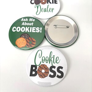 May include: Four round pin badges with cookie-themed designs. One badge says "Cookie Dealer" with a cookie graphic. Another says "Ask Me About COOKIES!" with cookie graphics. The third says "Cookie BOSS" with a cookie graphic. One badge is blank.