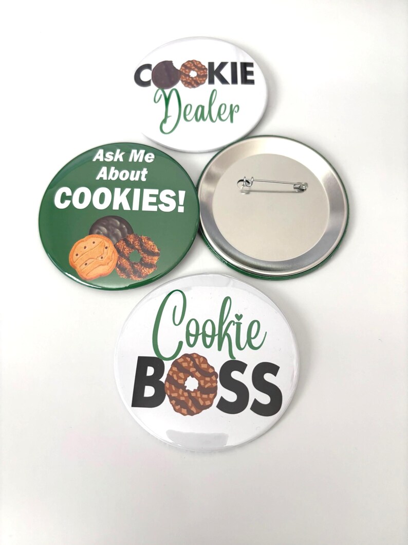 May include: Four round pin badges with cookie-themed designs. One badge says "Cookie Dealer" with a cookie graphic. Another says "Ask Me About COOKIES!" with cookie graphics. The third says "Cookie BOSS" with a cookie graphic. One badge is blank.