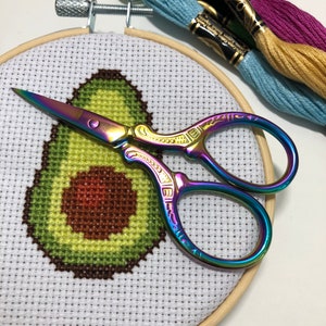 May include: A close-up of a completed cross-stitch embroidery project of an avocado on white Aida cloth in a wooden embroidery hoop. A pair of rainbow-colored embroidery scissors are lying on top of the hoop.