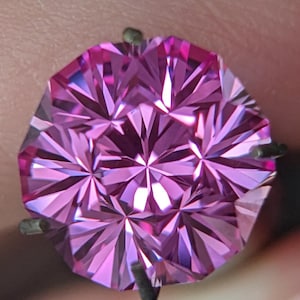May include: A faceted, round-cut, pink gemstone. The gem is held by a dark metal setting. The facets create a starburst pattern, reflecting light. The gem is held between fingers.