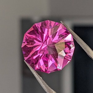 May include: A pink gemstone with a faceted cut, held by tweezers. The gemstone is a vibrant pink color and has a star-like pattern in the center.