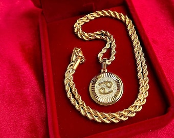 May include: A gold-coloured necklace with a pendant featuring the Cancer zodiac symbol, presented in a red velvet jewellery box. The necklace has a rope chain design. The pendant is round with a textured border.