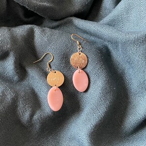 May include: A pair of earrings with a gold-colored, textured disc above a light pink oval. The earrings hang from silver-colored hooks. The background is a blue-gray fabric.