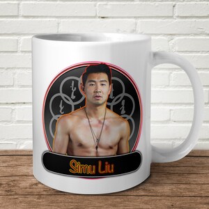 以下が含まれることがあります： 白いセラミック製のコーヒーカップ。黒と赤の円形デザインで、シム・リウの写真が描かれています。円形デザインは、赤い輪郭のある黒い背景に、連動する円の模様が描かれています。円の下の黒いバナーに白い文字で「シム・リウ」と印刷されています。