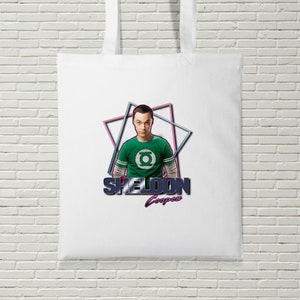 Puede incluir: Bolsa de tela blanca con un gráfico de Sheldon Cooper de la serie The Big Bang Theory. Lleva una camisa verde con un símbolo verde y el texto "SHELDON Cooper" está debajo.