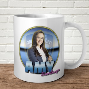 May include: White ceramic mug with a blue and yellow circle design featuring a woman with dark hair and a blue blazer. The text "AMY Santiago" is printed below the circle.