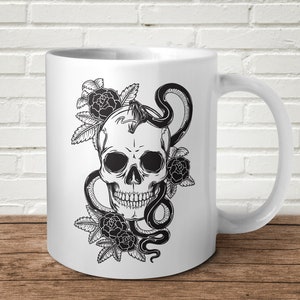 May include: White ceramic mug featuring a black and white illustration of a skull with a snake wrapped around it. The skull is surrounded by roses.