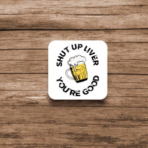 Puede incluir: Un posavasos cuadrado blanco con el texto "SHUT UP LIVER YOU'RE GOOD" en negro, que rodea una imagen de dibujos animados de una jarra de cerveza llena de cerveza amarilla y espuma blanca. El posavasos está sobre una superficie de madera marrón.