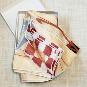 K&ouml;nnte beinhalten: Ein gefalteter Seidenschal mit cremefarbenem Hintergrund und einem farbenfrohen abstrakten Design aus roten, braunen und blauen Formen. Der Schal wird in einer wei&szlig;en Schachtel pr&auml;sentiert, was auf einen Geschenkartikel hindeutet. Die Kanten des Schals sind sauber verarbeitet.