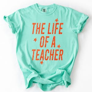 May include: A mint green t-shirt with the text "THE LIFE OF A TEACHER" in orange, with star accents. The shirt is made of a soft material and has rolled-up sleeves. The text is in a bold, textured font.