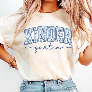 May include: Cream-colored t-shirt with the word "KINDER" in blue floral print lettering above the word "garten" in a cursive font. The shirt is a light, neutral color.