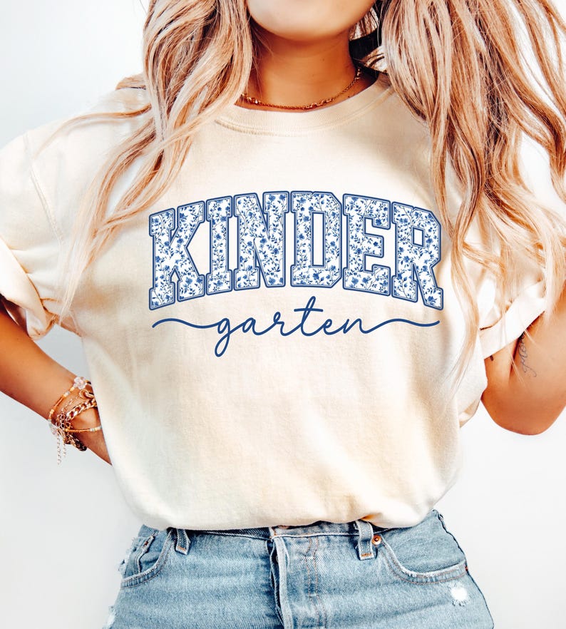 May include: Cream-colored t-shirt with the word "KINDER" in blue floral print lettering above the word "garten" in a cursive font. The shirt is a light, neutral color.