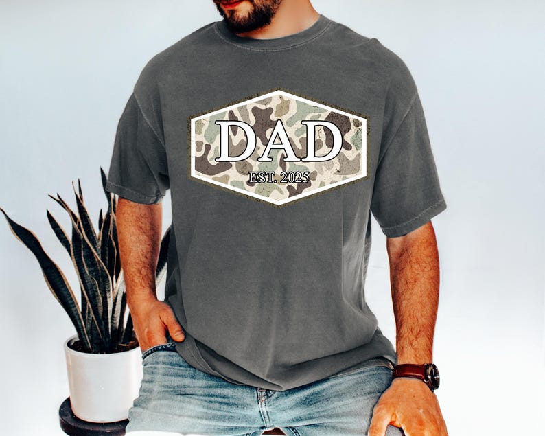 May include: A gray t-shirt with a camouflage design and the word "DAD" in white letters. The text "EST. 2025" is below the word "DAD". The shirt is a short-sleeved, casual style.