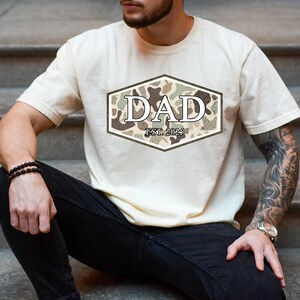 May include: Cream-colored t-shirt with a camouflage design in a hexagonal shape, featuring the word "DAD" and "EST. 2025". The shirt is a casual wear item.