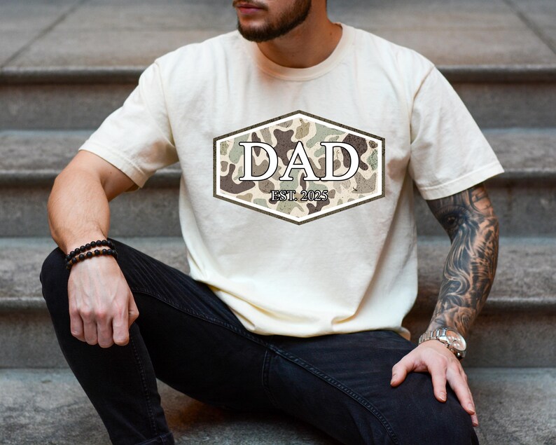 May include: Cream-colored t-shirt with a camouflage design in a hexagonal shape, featuring the word "DAD" and "EST. 2025". The shirt is a casual wear item.