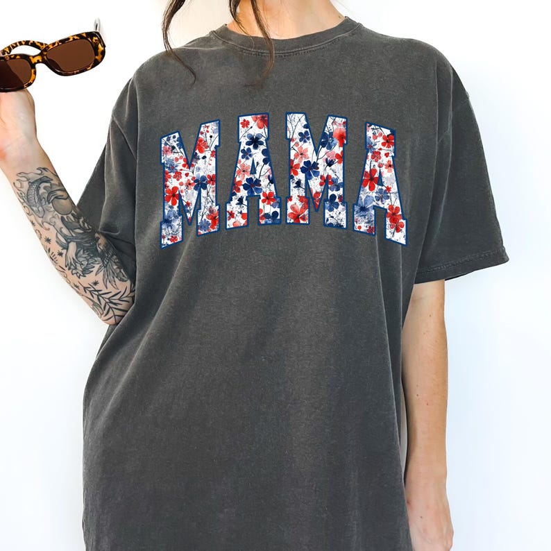 May include: Dark grey t-shirt with the word "MAMA" in a floral design with red, white, and blue flowers. The shirt has a vintage, washed look. Sunglasses are held in the upper left corner.