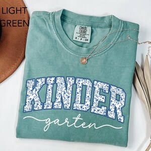 May include: Light green t-shirt with the word "KINDER" in blue floral print and "garten" in white script. A silver necklace with a pendant rests on the shirt. The shirt is folded.