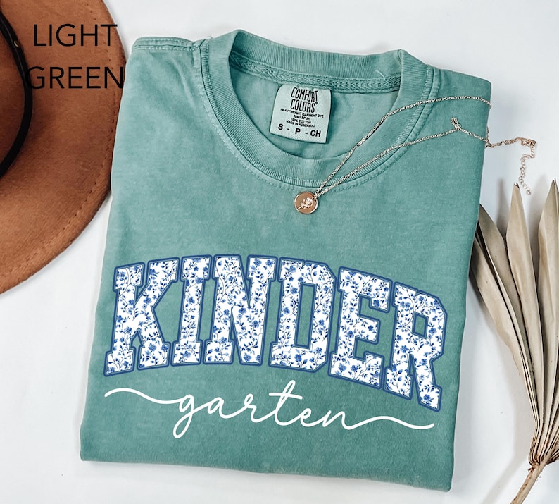 May include: Light green t-shirt with the word "KINDER" in blue floral print and "garten" in white script. A silver necklace with a pendant rests on the shirt. The shirt is folded.