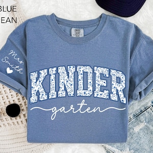May include: Blue jean-colored t-shirt with the word "KINDERGARTEN" in a floral design. The word "garten" is written in a cursive font. The sleeve has the text "Mrs. Smith" and a heart.