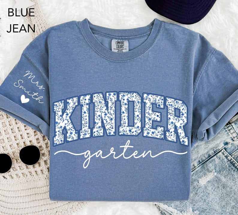 May include: Blue jean-colored t-shirt with the word "KINDERGARTEN" in a floral design. The word "garten" is written in a cursive font. The sleeve has the text "Mrs. Smith" and a heart.