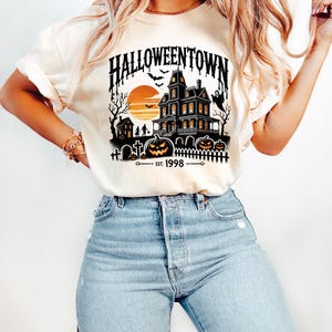 May include: A white t-shirt with a black graphic of a spooky house, pumpkins, and bats. The text "HALLOWEENTOWN EST. 1998" is printed on the shirt.