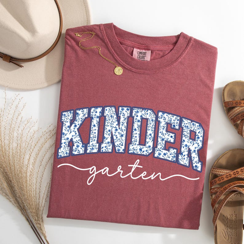 May include: A maroon Comfort Colors t-shirt with the word "KINDER" in blue floral print and "garten" in white script. A gold necklace and a beige hat are next to the shirt. Brown sandals are on the right.