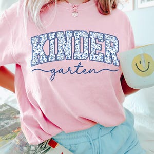 May include: A light pink t-shirt with the word "KINDER" in a blue floral design and the word "garten" in a cursive font. A white mug with a yellow smiley face is held in the hand.