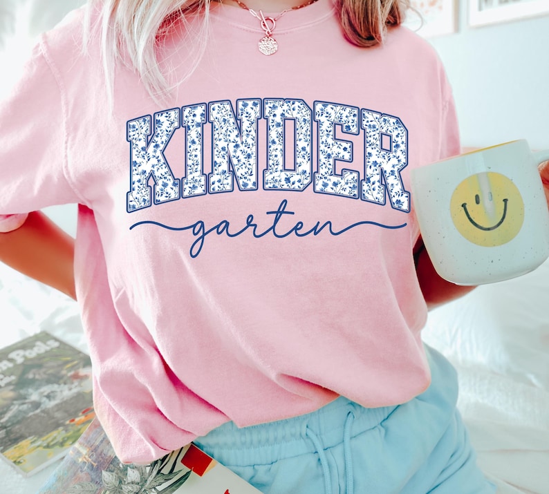 May include: A light pink t-shirt with the word "KINDER" in a blue floral design and the word "garten" in a cursive font. A white mug with a yellow smiley face is held in the hand.