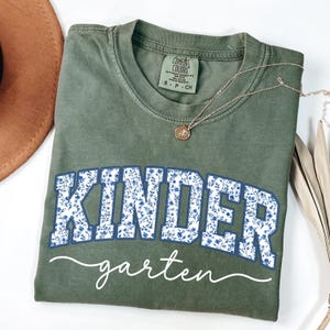 May include: An olive green t-shirt with the word "KINDER" in large, blue floral letters, and "garten" in white script. A gold necklace with a pendant is draped across the shirt. The shirt is a Comfort Colors brand.
