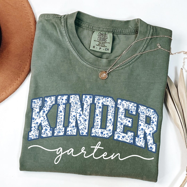 May include: An olive green t-shirt with the word "KINDER" in large, blue floral letters, and "garten" in white script. A gold necklace with a pendant is draped across the shirt. The shirt is a Comfort Colors brand.