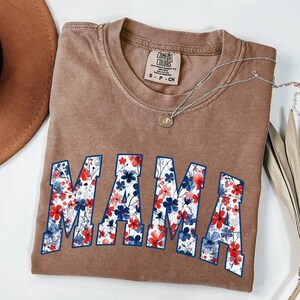 May include: A brown Comfort Colors t-shirt with the word "MAMA" in a floral design. The letters are filled with red and blue flowers. A silver necklace with a pendant is draped across the shirt.