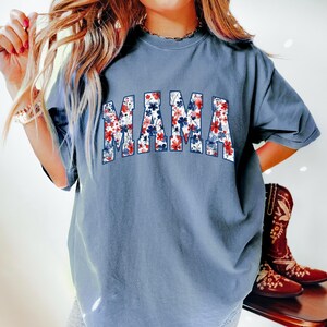 May include: A dusty blue t-shirt with the word "MAMA" in a floral design. The letters are filled with red, white, and blue flowers. The shirt has short sleeves and a relaxed fit. The image also shows a pair of brown cowboy boots.